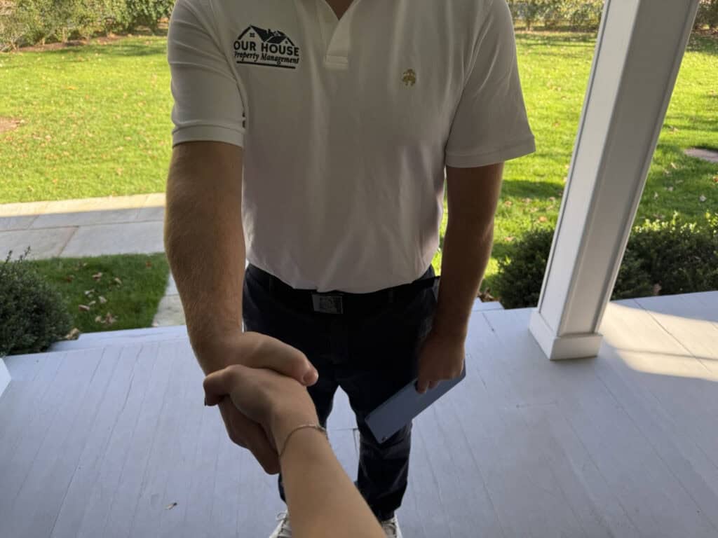 A person in a white polo shirt is shaking hands with someone on a doorstep. The shirt has a small logo with text that reads "Our House" and "moving." The person holds a clipboard or tablet in the other hand. A grassy yard is visible in the background.