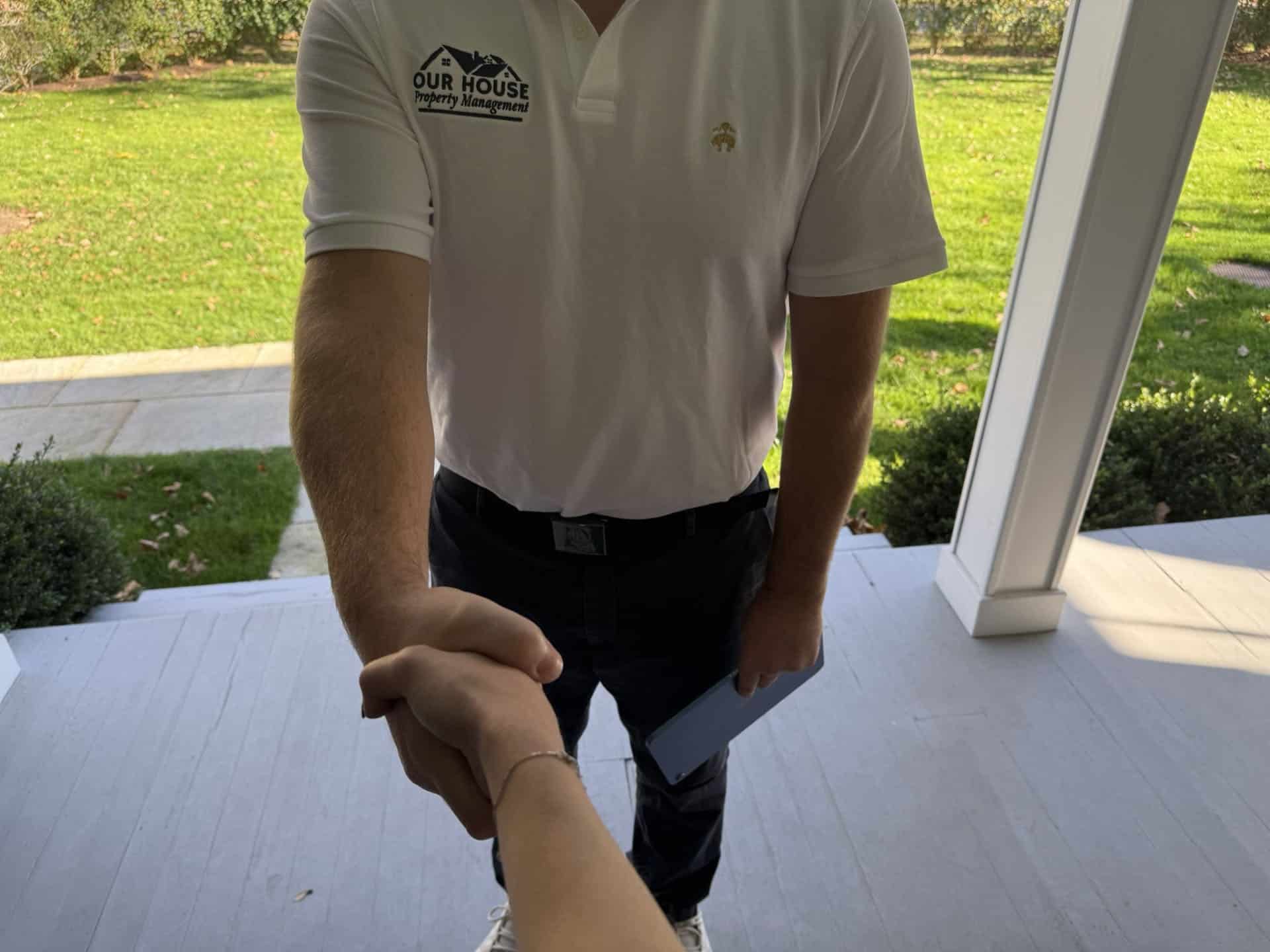 A person in a white polo shirt is shaking hands with someone on a doorstep. The shirt has a small logo with text that reads "Our House" and "moving." The person holds a clipboard or tablet in the other hand. A grassy yard is visible in the background.