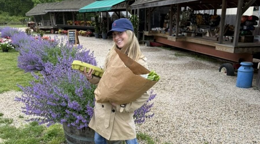 A smiling woman in a tan coat, blue cap, and jeans holds a large paper bag of produce at an outdoor market, standing near purple flowers with wooden stalls and greenery in the background.