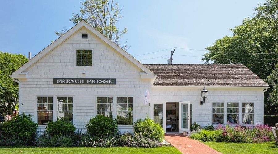 A white, cottage-style building with a sign reading "French Presse" above the main entrance, surrounded by greenery, flowers, and a brick walkway under a clear blue sky.
