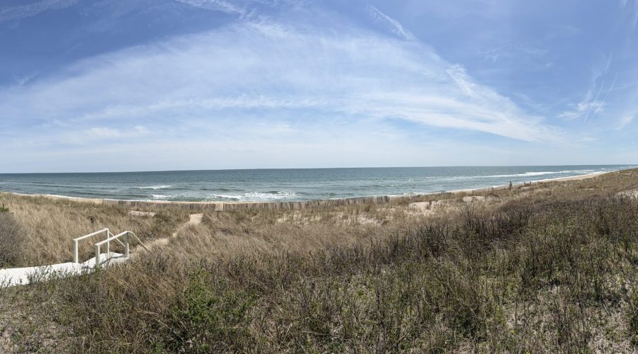 A panoramic view of a sandy beach with grassy dunes, a pathway and steps leading to the ocean, under a blue sky with wispy clouds.
