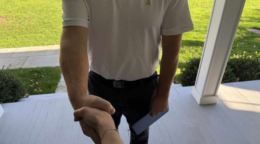 A person in a white polo shirt is shaking hands with someone on a doorstep. The shirt has a small logo with text that reads "Our House" and "moving." The person holds a clipboard or tablet in the other hand. A grassy yard is visible in the background.