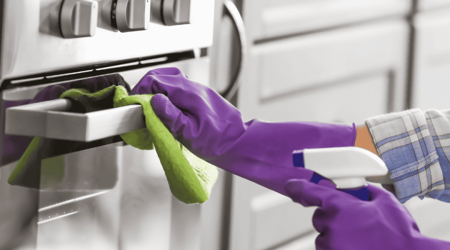 A person wearing purple gloves cleans a stainless steel kitchen appliance with a green cloth while spraying cleaner. The background shows modern kitchen cabinets and drawers with a sleek, shiny finish, exemplifying the meticulous care provided by Property Management East Hampton.