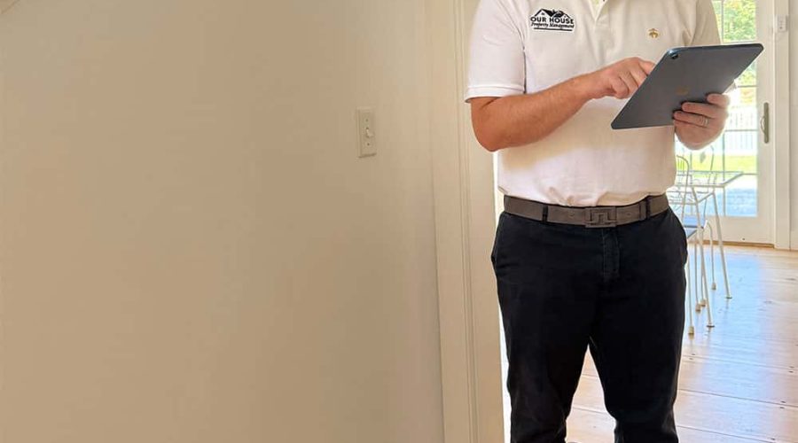 A man wearing a white polo shirt and black pants stands in a room with wooden flooring, using a tablet. He has blue shoe covers and a black cap. The background features a window and part of a chair.