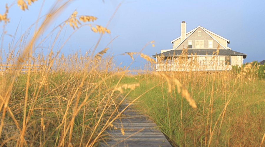 A wooden pathway leads through tall grass towards a two-story Hamptons beach house with a green roof. The house, surrounded by golden reeds, sits against a clear blue sky, evoking a serene, coastal atmosphere. Managed by Luxury Home Management East Hampton, this image suggests a peaceful seaside retreat.