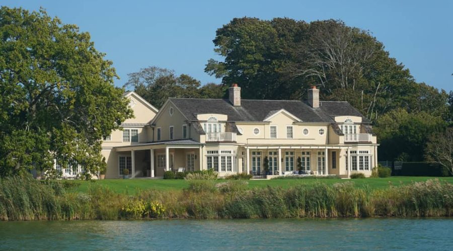 A large, elegant house with multiple chimneys and expansive windows sits near a calm body of water. Managed by a top-tier rental property management team, the property is surrounded by lush green trees and grass under a clear blue sky, ensuring both beauty and peace for its residents.